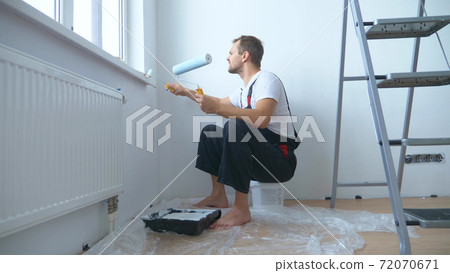 handsome man in work overalls paints the wall with a roller white paint handsome man in work overalls paints the wall with a roller white paint 72070671