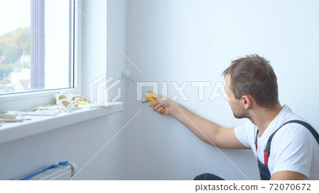 handsome man in work overalls paints the wall with a roller white paint 72070672