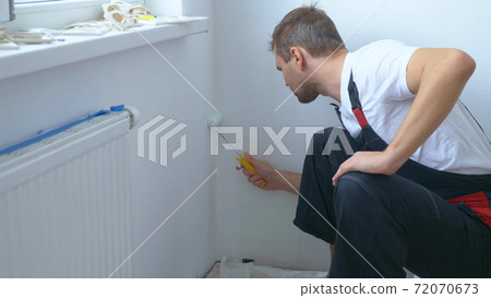 handsome man in work overalls paints the wall with a roller white paint handsome man in work overalls paints the wall with a roller white paint 72070673