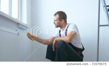 handsome man in work overalls paints the wall with a roller white paint handsome man in work overalls paints the wall with a roller white paint 72070674
