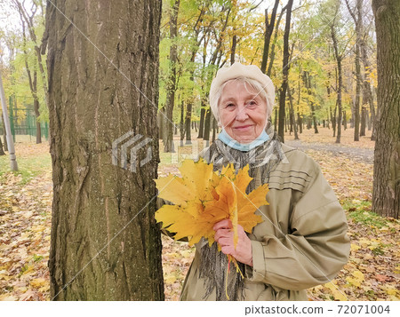 Older woman in protective mask and a bouquet of autumn leaves is having fun in the autumn park outdoors. Older woman in protective mask and a bouquet of autumn leaves is having fun in the autumn park outdoors. 72071004