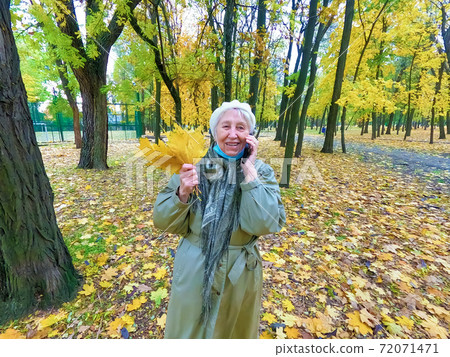 Older woman in protective mask and a bouquet of autumn leaves is having fun in the autumn park outdoors. Older woman in protective mask and a bouquet of autumn leaves is having fun in the autumn park outdoors. 72071471