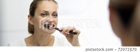 Woman in awhite T-shirt is brushing her teeth in front of mirror 72072870