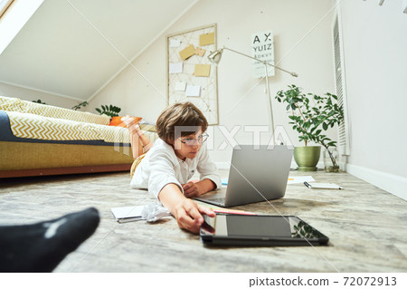 Homeschooling. Small boy wearing eyeglasses lying on the wooden floor in the living room and studying online, he is using laptop and digital tablet while doing homework 72072913