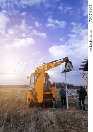 The excavator with a bucket pushes the pillars on the fence in the field near the house 72074161