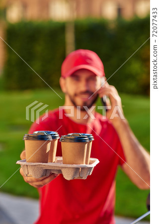 Young caucasian male courier talking by smartphone while delivering four coffee cups to custumer Young caucasian male courier talking by smartphone while delivering four coffee cups to custumer 72077433