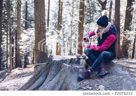 A smiling woman tourist pours hot tea from a thermos into a metal mug while sitting on a tree stump. A smiling woman tourist pours hot tea from a thermos into a metal mug while sitting on a tree stump. 72078232