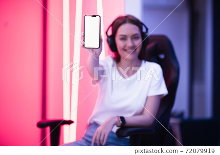 Smiling gamer girl wearing headset showing smart phone and pointing at blank screen with her finger. Smiling gamer girl wearing headset showing smart phone and pointing at blank screen with her finger. 72079919