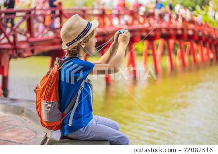 Caucasian boy tourist on background of Red Bridge in public park garden with trees and reflection in the middle of Hoan Kiem Lake in Downtown Hanoi. Traveling with children concept 72082316