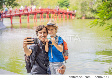 Caucasian Dad and son travelers on background of Red Bridge in public park garden with trees and reflection in the middle of Hoan Kiem Lake in Downtown Hanoi. Traveling with children concept 72082347