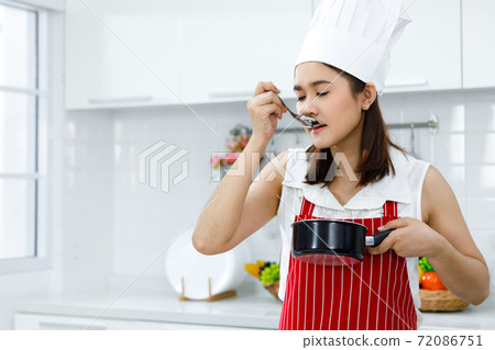 Asian woman chef in red apron taste soup from pot. Concept woman preparing meals at home. Asian woman chef in red apron taste soup from pot. Concept woman preparing meals at home. 72086751