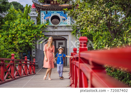 Caucasian mom and son travelers on background of Red Bridge in public park garden with trees and reflection in the middle of Hoan Kiem Lake in Downtown Hanoi. Traveling with children concept 72087402