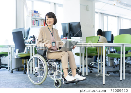Portrait of a woman in a wheelchair working with her laptop on her lap in the office 72089420