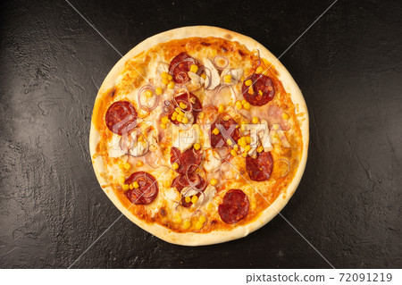 Round hot freshly baked pizza with salami, chicken, corn, onion and cheese lies on a black stone kitchen table Round hot freshly baked pizza with salami, chicken, corn, onion and cheese lies on a black stone kitchen table 72091219
