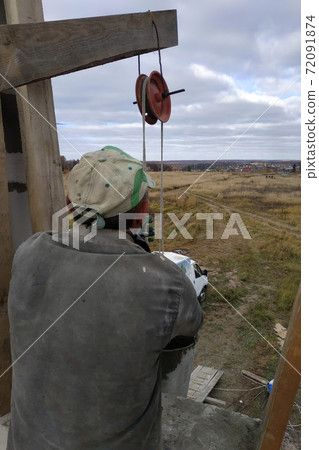 A worker using a hand winch raises a cement mortar bin on the second floor A worker using a hand winch raises a cement mortar bin on the second floor 72091874