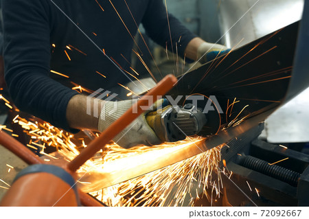 Locksmith in special clothes and goggles works in production. Metal processing with angle grinder. Sparks in metalworking 72092667