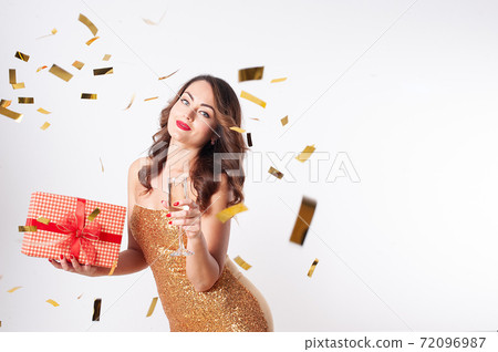 Young woman holding a gift box and drinks champagne, having fun with golden confetti 72096987