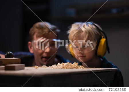 an experienced carpenter and a small boy in safety glasses blow sawdust off a work table in a carpenter's workshop. 72102388