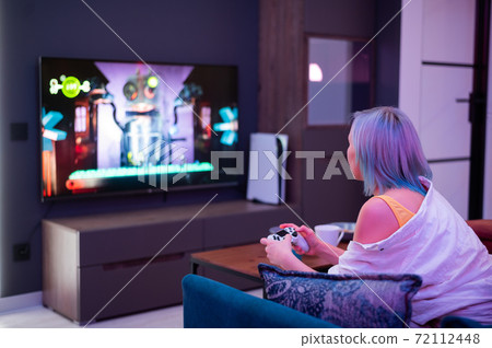 Teenager girl with colorful blue and pink hair hold joystick and play video games at home at night. 72112448