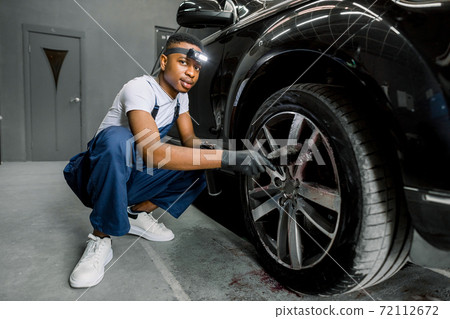 Professional African male worker in overalls and black protective gloves, cleaning alloy wheels rims of luxury car with a special brush for cast wheels in a detailing workshop, looking at camera Professional African male worker in overalls and black protective gloves, cleaning alloy wheels rims of luxury car with a special brush for cast wheels in a detailing workshop, looking at camera 72112672