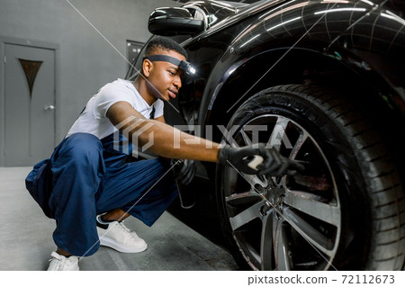 Washing a car by hand, car detailing. Close up image of professional African male worker, cleaning the car wheels with brush. Car rims wash, manual wheel cleaning concept. Washing a car by hand, car detailing. Close up image of professional African male worker, cleaning the car wheels with brush. Car rims wash, manual wheel cleaning concept. 72112673
