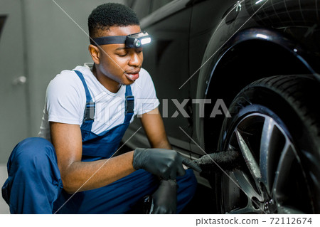 Car washing and detailing workshop. African man worker in protective overalls and rubber gloves, washing car alloy wheel on a car wash, using special brush. Close up portrait, selective focus Car washing and detailing workshop. African man worker in protective overalls and rubber gloves, washing car alloy wheel on a car wash, using special brush. Close up portrait, selective focus 72112674