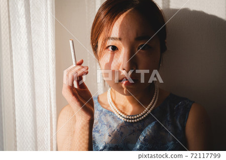 在房間的窗戶旁拿著煙的藍色連衣裙的年輕女子 72117799