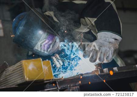 Blacksmith welder in protective mask works with metal steel and iron using welding machine, bright sparks and flashes. Welder uses mask to protect eyes from spark. Professional industry welder working Blacksmith welder in protective mask works with metal steel and iron using welding machine, bright sparks and flashes. Welder uses mask to protect eyes from spark. Professional industry welder working 72123817