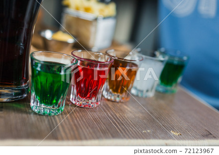 Multi-color cool alcohol party drinks with on wooden table in bar. Bright colorful glasses with alcoholic shots. Mixed liquor, vodka and juice in shot glasses. Alcoholic cocktail row on bar table. 72123967