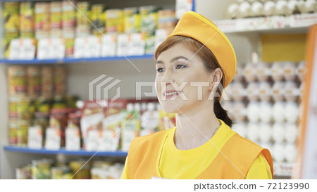Portrait of smiling Caucasian female staff standing in grocery section of supermarket. Cheerful slender saleswoman in yellow t-shirt andorange apron. Trade business and people concept Portrait of smiling Caucasian female staff standing in grocery section of supermarket. Cheerful slender saleswoman in yellow t-shirt andorange apron. Trade business and people concept 72123990