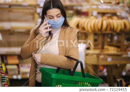 Buyer wearing a protective mask and gloves in supermarket. Shopping during the pandemic quarantine Covid-19. Nonperishable smart purchased household pantry groceries preparation. Woman spear by phone Buyer wearing a protective mask and gloves in supermarket. Shopping during the pandemic quarantine Covid-19. Nonperishable smart purchased household pantry groceries preparation. Woman spear by phone 72129455