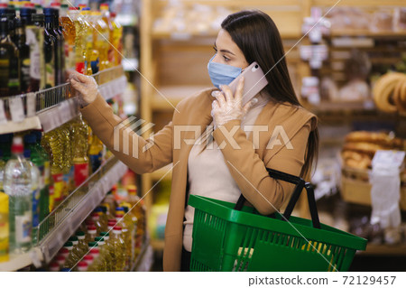 Buyer wearing a protective mask and gloves in supermarket. Shopping during the pandemic quarantine Covid-19. Nonperishable smart purchased household pantry groceries preparation. Woman spear by phone Buyer wearing a protective mask and gloves in supermarket. Shopping during the pandemic quarantine Covid-19. Nonperishable smart purchased household pantry groceries preparation. Woman spear by phone 72129457