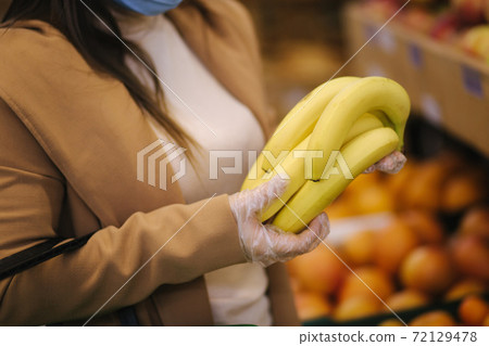 Close up of Young woman in protective gloves and face mask hold beautiful fresh bananas in hand. Beautiful young girl with food basket choosing food by stand with fruits. Shopping during quarantine Close up of Young woman in protective gloves and face mask hold beautiful fresh bananas in hand. Beautiful young girl with food basket choosing food by stand with fruits. Shopping during quarantine 72129478