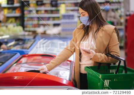 Female shopping during the coronavirus Covid-19 pandemic. A young woman buys semi finished food from refregerator in a supermarket with shopping cart. Woman in facial mask and gloves to prevent Female shopping during the coronavirus Covid-19 pandemic. A young woman buys semi finished food from refregerator in a supermarket with shopping cart. Woman in facial mask and gloves to prevent 72129490