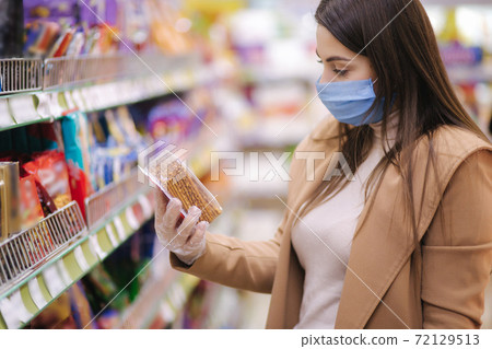 Horizontal shot of woman in protective mask and gloves reading label with price at goods while standing in supermarket. Beautiful young girl choosing snaks while doing shopping at grocery shop Horizontal shot of woman in protective mask and gloves reading label with price at goods while standing in supermarket. Beautiful young girl choosing snaks while doing shopping at grocery shop 72129513
