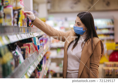 Horizontal shot of woman in protective mask and gloves reading label with price at goods while standing in supermarket. Beautiful young girl choosing snaks while doing shopping at grocery shop Horizontal shot of woman in protective mask and gloves reading label with price at goods while standing in supermarket. Beautiful young girl choosing snaks while doing shopping at grocery shop 72129517