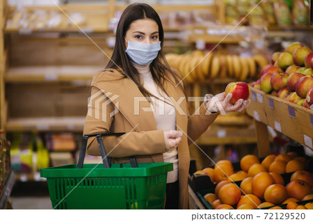 Young woman in protective gloves and face mask hold beautiful fresh apple in hand. Beautiful young girl with food basket choosing food by stand with fruits. Shopping during quarantine. Covid-19 Young woman in protective gloves and face mask hold beautiful fresh apple in hand. Beautiful young girl with food basket choosing food by stand with fruits. Shopping during quarantine. Covid-19 72129520