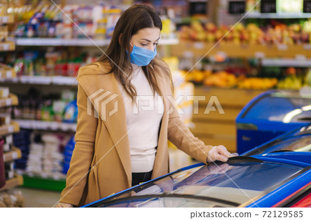 Female shopping during the coronavirus Covid-19 pandemic. A young woman buys semi finished food from refregerator in a supermarket with shopping cart. Woman in facial mask and gloves to prevent Female shopping during the coronavirus Covid-19 pandemic. A young woman buys semi finished food from refregerator in a supermarket with shopping cart. Woman in facial mask and gloves to prevent 72129585