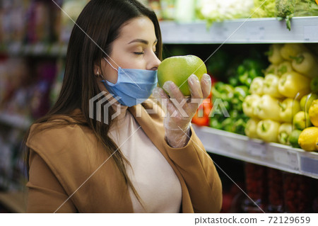 Happy woman in protective mask taking fresh vegetables while standing by groceries in supermarket. Beautiful young girl with food basket choosing food by stand with Fruits. Woman smell pomelo Happy woman in protective mask taking fresh vegetables while standing by groceries in supermarket. Beautiful young girl with food basket choosing food by stand with Fruits. Woman smell pomelo 72129659