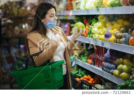 Happy woman in protective mask taking fresh vegetables while standing by groceries in supermarket. Beautiful young girl with food basket choosing food by stand with Fruits. Christmas purchases Happy woman in protective mask taking fresh vegetables while standing by groceries in supermarket. Beautiful young girl with food basket choosing food by stand with Fruits. Christmas purchases 72129661