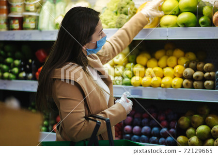 Happy woman in protective mask taking fresh vegetables while standing by groceries in supermarket. Beautiful young girl with food basket choosing food by stand with vegetables. Christmas purchases Happy woman in protective mask taking fresh vegetables while standing by groceries in supermarket. Beautiful young girl with food basket choosing food by stand with vegetables. Christmas purchases 72129665