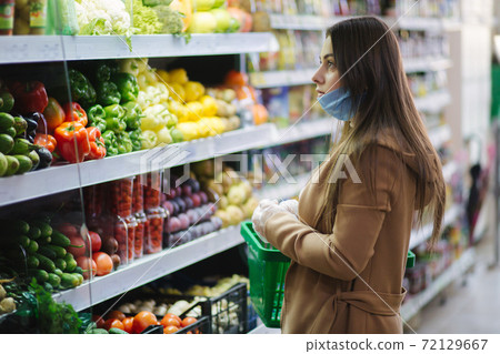Happy woman in protective mask taking fresh vegetables while standing by groceries in supermarket. Beautiful young girl with food basket choosing food by stand with vegetables. Christmas purchases 72129667
