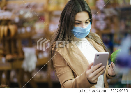 Happy woman in face mask hold phone and choose vegetables in supermarket. Beautiful young business woman choosing groceries by stand with vegetables. Quarantine. Covid-19 Happy woman in face mask hold phone and choose vegetables in supermarket. Beautiful young business woman choosing groceries by stand with vegetables. Quarantine. Covid-19 72129683
