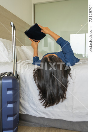 Woman lying on bed using tablet next to carry-on luggage 72129704