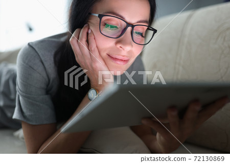 Young woman with glasses is lying on couch and looking into tablet at home 72130869