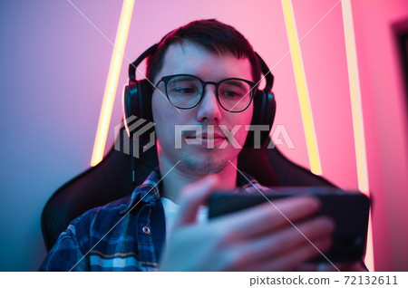 Close up portrait of a boy relaxing playing games on mobile phone and sitting in a gaming chair. Room with colorful neon light 72132611