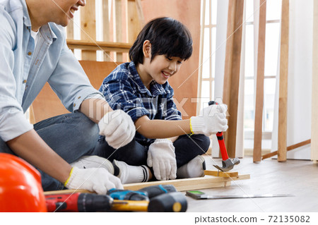 Asian boy sitting beside his father and using hammer to remove nail from timber. Concept family doing hobby together. 72135082