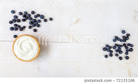 Heap of juicy blueberries and wooden bowl of smooth white yoghurt on white table Heap of juicy blueberries and wooden bowl of smooth white yoghurt on white table 72135186