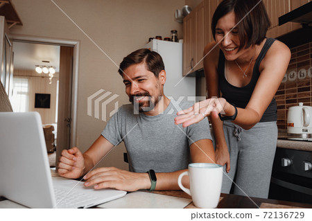 Couple at kitchen, reading news and using laptop. Couple at kitchen, reading news and using laptop. 72136729