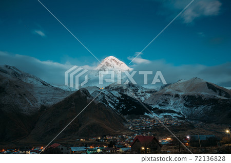 Stepantsminda, Georgia. Peak Of Mount Kazbek Covered With Snow And Famous Gergeti Church In Early Morning Lightning. Beautiful Georgian Landscape In Early Winter 72136828